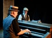 Two older men dressed in vintage-style clothing play music, one on piano in the foreground, the other smiling and playing drums in the background.