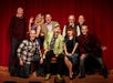 A group of nine adults, some seated and some standing, pose smiling in front of a red theater curtain.