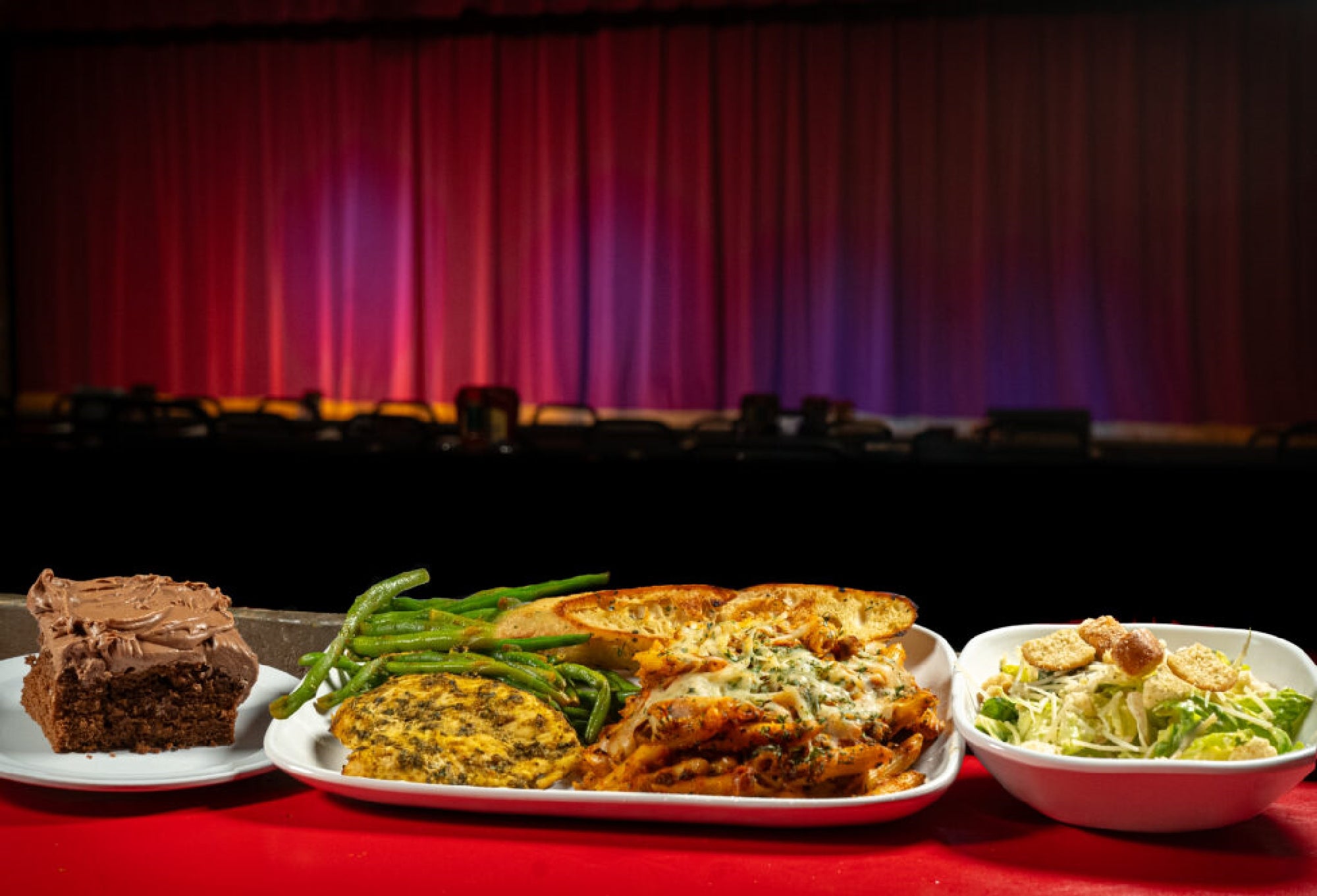 A plate with green beans, a breaded patty, cheesy pasta, and garlic bread, alongside a salad and a piece of chocolate cake, set in front of a stage with a closed red curtain.