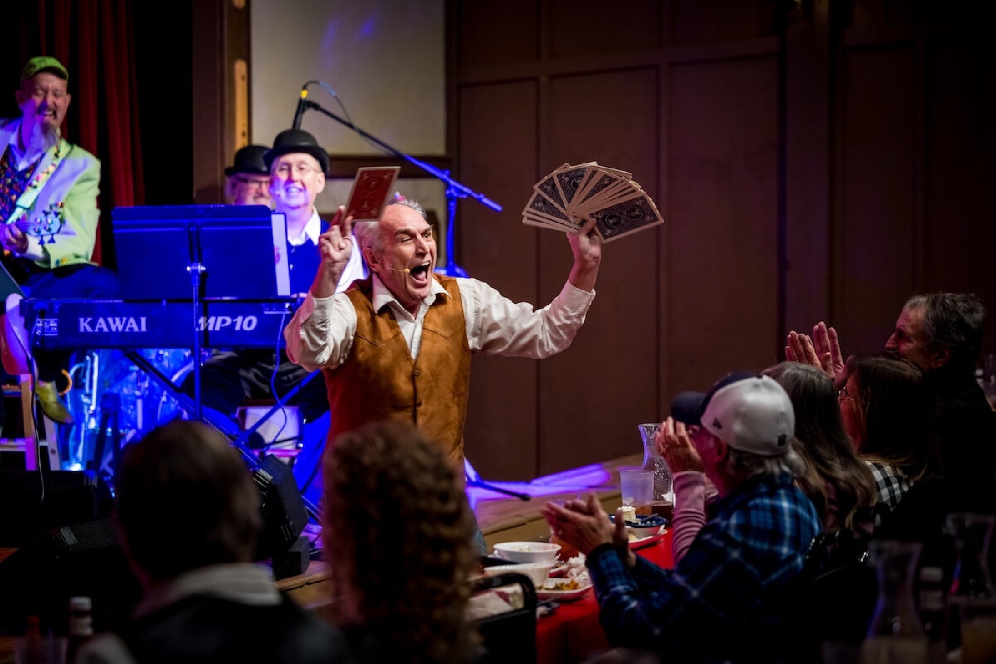 A performer on stage holds up oversized fake dollar bills and a card, entertaining an applauding audience at a dinner show with musicians in the background.