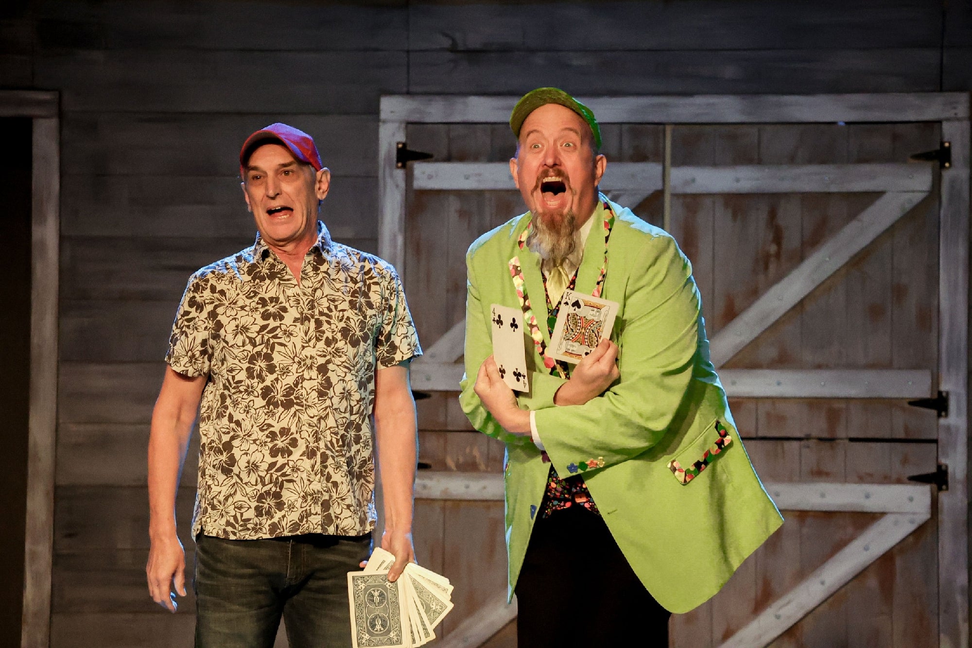 Two men perform on stage; one in a floral shirt and red cap holding cards, the other in a green jacket excitedly displaying playing cards, with wooden barn doors in the background.