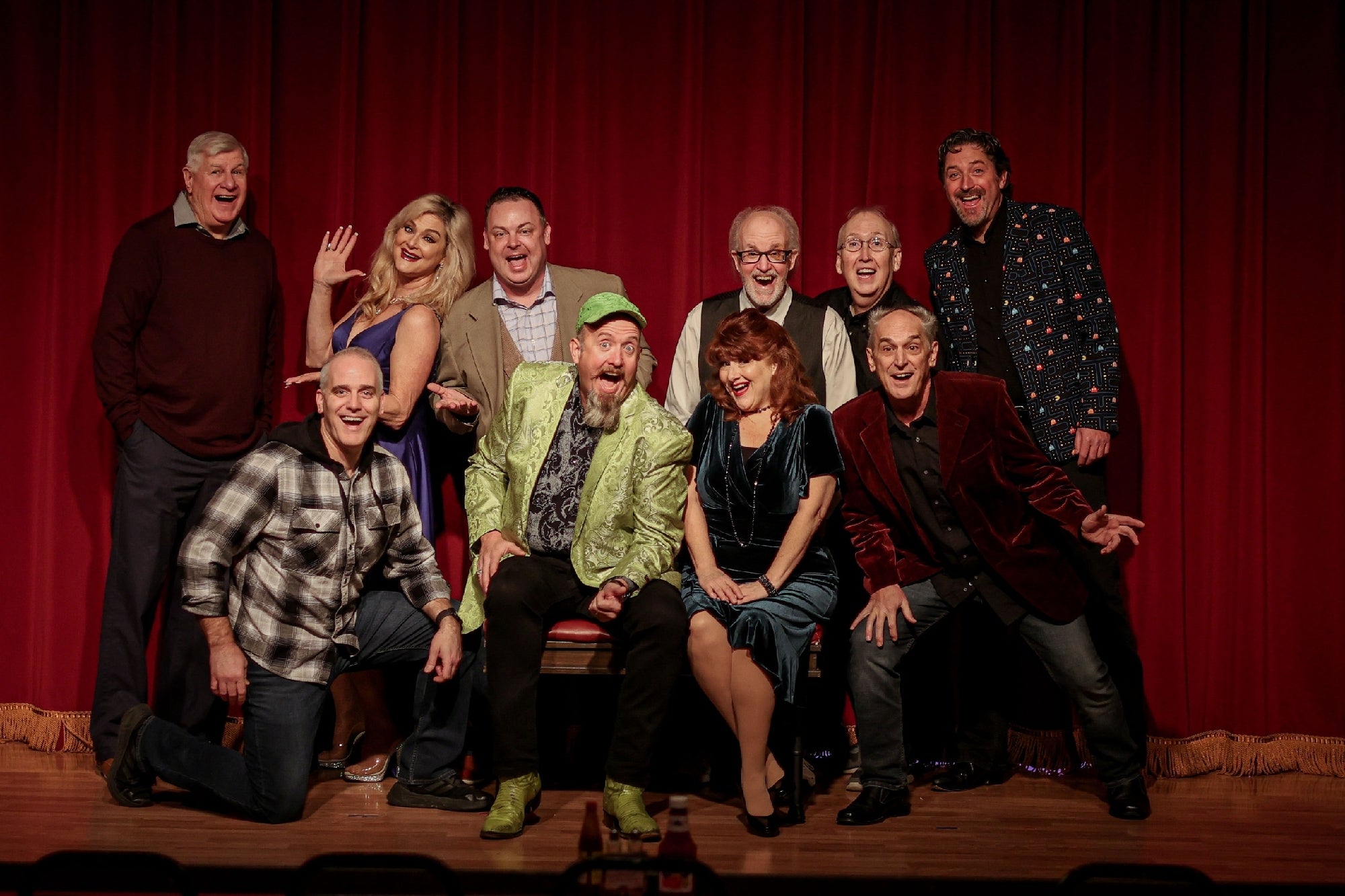 A group of ten people pose enthusiastically in front of a red curtain, with some seated and others standing behind.