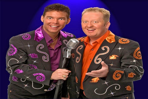 Two men in colorful, embellished suits stand together, one holding a vintage microphone, against a dark background.