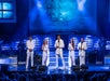 Three men in white suits and two women in white dresses performing on stage with blue lights shining behind them.