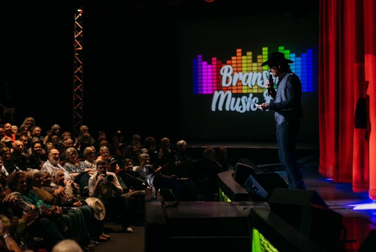 Performer in a cowboy hat stands on stage speaking to a seated audience, with a colorful "Branson Music" graphic projected behind him.