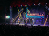Performers on stage at nighttime under colorful lights with a "Nashville Roadhouse Live" sign in the background and an audience watching.
