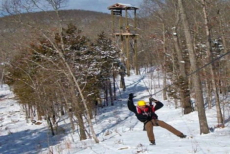 Person wearing a yellow helmet and harness ziplining through a snowy, wooded area with a wooden observation tower in the background.