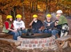 Six people wearing helmets sit around a campfire pit in a wooded area during autumn, holding drinks and smiling at the camera.