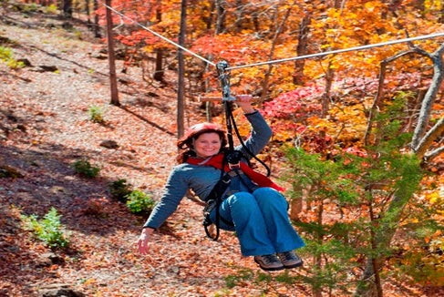 A woman wearing a harness rides a zipline through a forest with colorful autumn foliage.