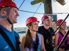 Four people wearing helmets and harnesses stand outdoors, smiling and looking ahead, likely preparing for a ziplining or ropes course activity.