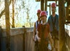 Three people wearing helmets and harnesses walk along a wooden and metal walkway outdoors, with trees and sunlight in the background.