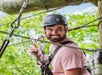 A man wearing a helmet and harness smiles while participating in a high ropes course outdoors.