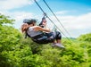 A person wearing a helmet and harness rides a zip line over a green forest on a sunny day.
