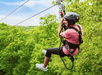 A person wearing a helmet and harness glides on a zip line above green trees on a sunny day.