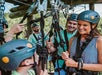 A group of people wearing helmets and harnesses prepare for a ropes course outdoors, smiling and interacting under a wooden structure.