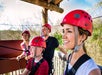Four people wearing red helmets and harnesses stand on a wooden platform, smiling and preparing for a zipline activity outdoors.