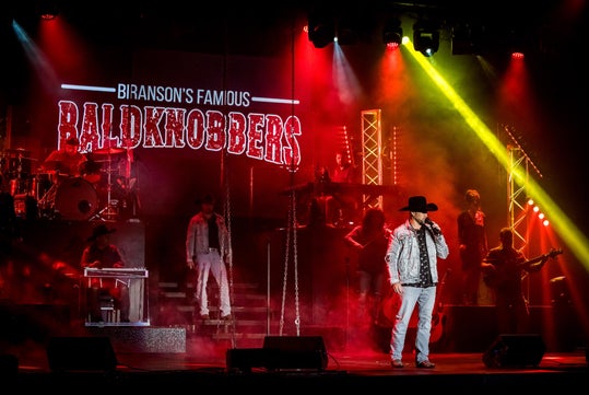 A band performs on a stage with "Branson's Famous Baldknobbers" displayed in lights behind them, with red and yellow stage lighting and smoke effects.