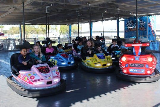 People of various ages are riding bumper cars in an indoor amusement park attraction, with several cars visible and the area lit by daylight.