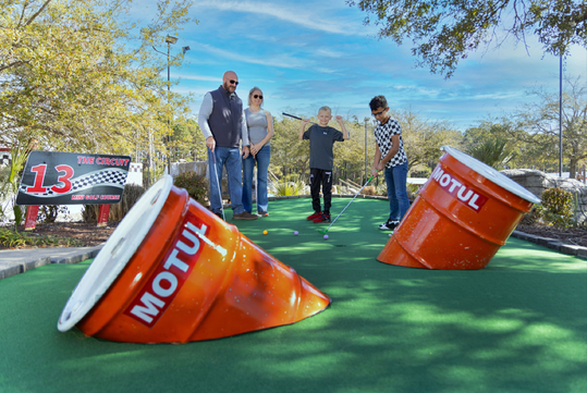 People playing mini-golf on a course with oversized bucket obstacles. A child is aiming to putt while others watch.
