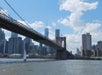 The Brooklyn Bridge spans over the East River with a sailboat below and the Manhattan skyline in the background on a partly cloudy day.
