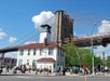 A crowd of people walk past a white building near the base of the Brooklyn Bridge on a sunny day with blue sky and clouds.