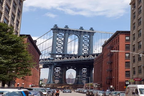 View of the Manhattan Bridge framed by red brick buildings in Brooklyn, with the Empire State Building visible through the bridge’s arch. Cars are parked along the street.