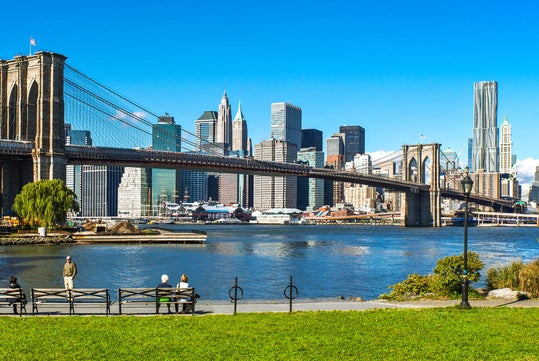 View of the Brooklyn Bridge with New York City skyline in the background on a clear day. People are sitting on benches in a park by the water.