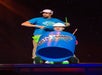 Two men perform on stage; one stands behind playing drumsticks, while the other sits inside a large blue plastic barrel wearing a hard hat.