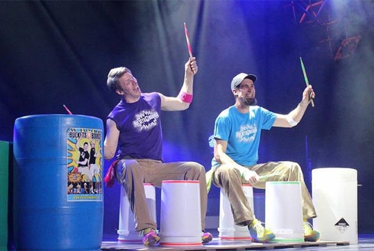 Two performers sit on plastic buckets on stage, wearing colorful t-shirts and holding drumsticks, surrounded by large barrels and stage lights.