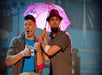 Two men on stage share a pink Hello Kitty umbrella; one gives a thumbs up while the other looks apprehensive. Both wear casual clothing and colorful wristbands.