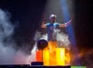 A person wearing a cap and wristbands plays drums made from large barrels on a smoky, dimly lit stage.