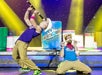 Two performers in colorful outfits play tubes on stage, with a large barrel labeled "Buckets N Boards" and drum props in the background.