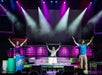 Three performers on stage raise their arms, each standing behind colorful drums, with bright spotlights and a vibrant purple and green backdrop.