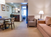Hotel suite interior with a small dining table, chairs, sofa, and lamp; kitchenette and bedroom visible through an open door.