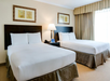 Hotel room with two neatly made double beds, a bedside table with a lamp, two framed pictures above the beds, and a window with curtains.
