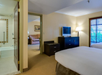 Hotel room with a bed, TV, and dresser. Bathroom with a tiled bathtub visible to the left. An adjacent room with a sofa can be seen through the open door.