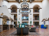 Spacious hotel lobby with a large chandelier, seating area, and decorative plants, surrounded by multiple balconies and arches.