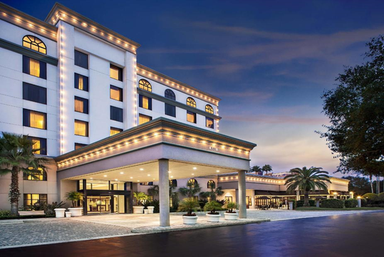 A multi-story hotel with illuminated exterior lights, a covered entrance, palm trees, and a paved driveway at dusk.