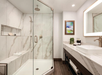 Modern bathroom with a glass-enclosed shower, marble walls, a large vanity with a vessel sink, wall mirror, and a colorful framed painting. Towels and toiletries are neatly arranged.