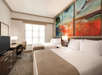 Hotel room with two double beds, a desk with a chair, a flat-screen TV, and large abstract artwork above the beds. Natural light enters through a window with blinds.