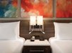 Two beds with white linens flank a nightstand holding a phone and clock, beneath modern wall lamps and colorful abstract paintings.