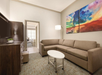 A modern hotel suite living area with a beige sectional sofa, a round white coffee table, wall-mounted TV, and colorful abstract artwork above the couch. An adjacent room is visible.