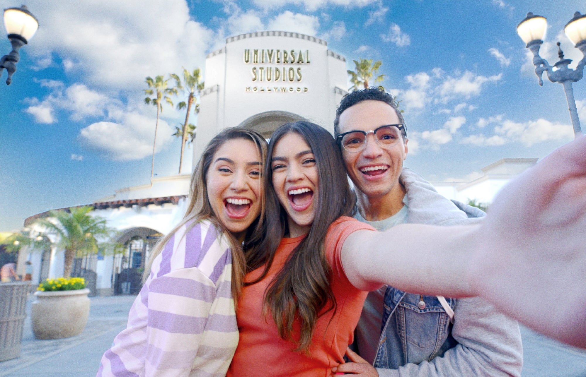 Three young adults smiling and taking a selfie in front of the Universal Studios Hollywood entrance on a sunny day.