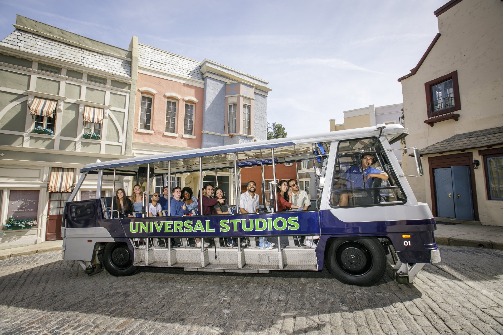 A Universal Studios tour tram filled with passengers drives through a themed set with colorful buildings on a cobblestone street.