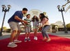 A family of four smiles and laughs together while standing on a red carpet in front of the Universal Studios entrance on a sunny day.
