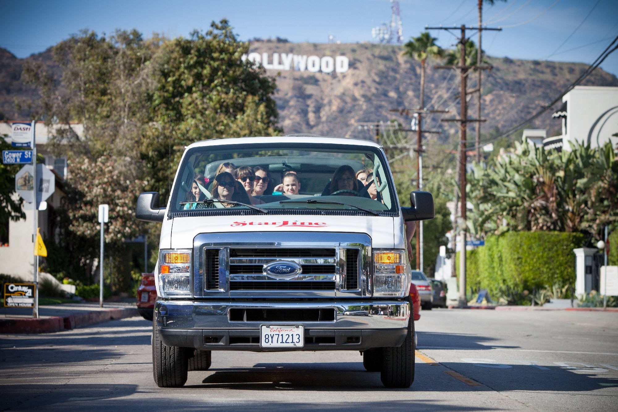 A white tour van with passengers drives on a street in Los Angeles, with the Hollywood sign visible on the hillside in the background.