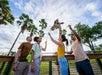 Four people stand on a platform feeding a giraffe at a zoo, with palm trees and a partly cloudy sky in the background.