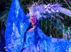 A performer dressed in an elaborate blue costume with sparkling details and a decorative headpiece stands with arms outstretched against a backdrop of greenery.