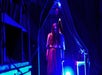 A person with long hair, wearing an old-fashioned dress, stands under blue lighting between dark curtains in a dimly lit room.