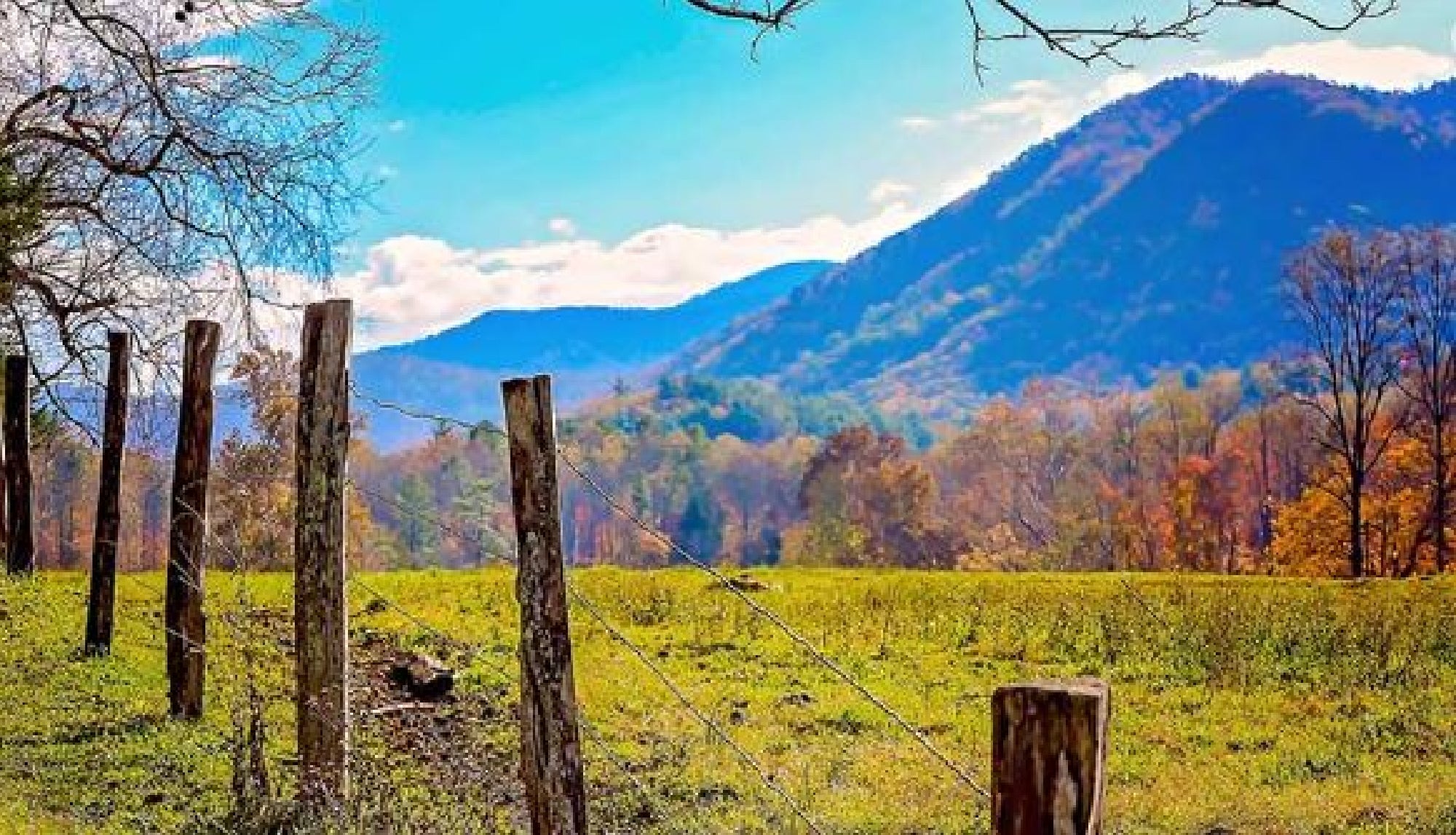 A rustic wooden fence in a grassy field, with trees showcasing autumn foliage. In the background, a range of blue mountains under a partly cloudy sky.